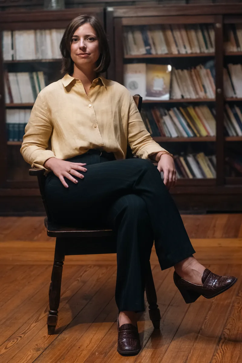 Juliana Vasconcelos Senra sentada numa cadeira de madeira, numa biblioteca